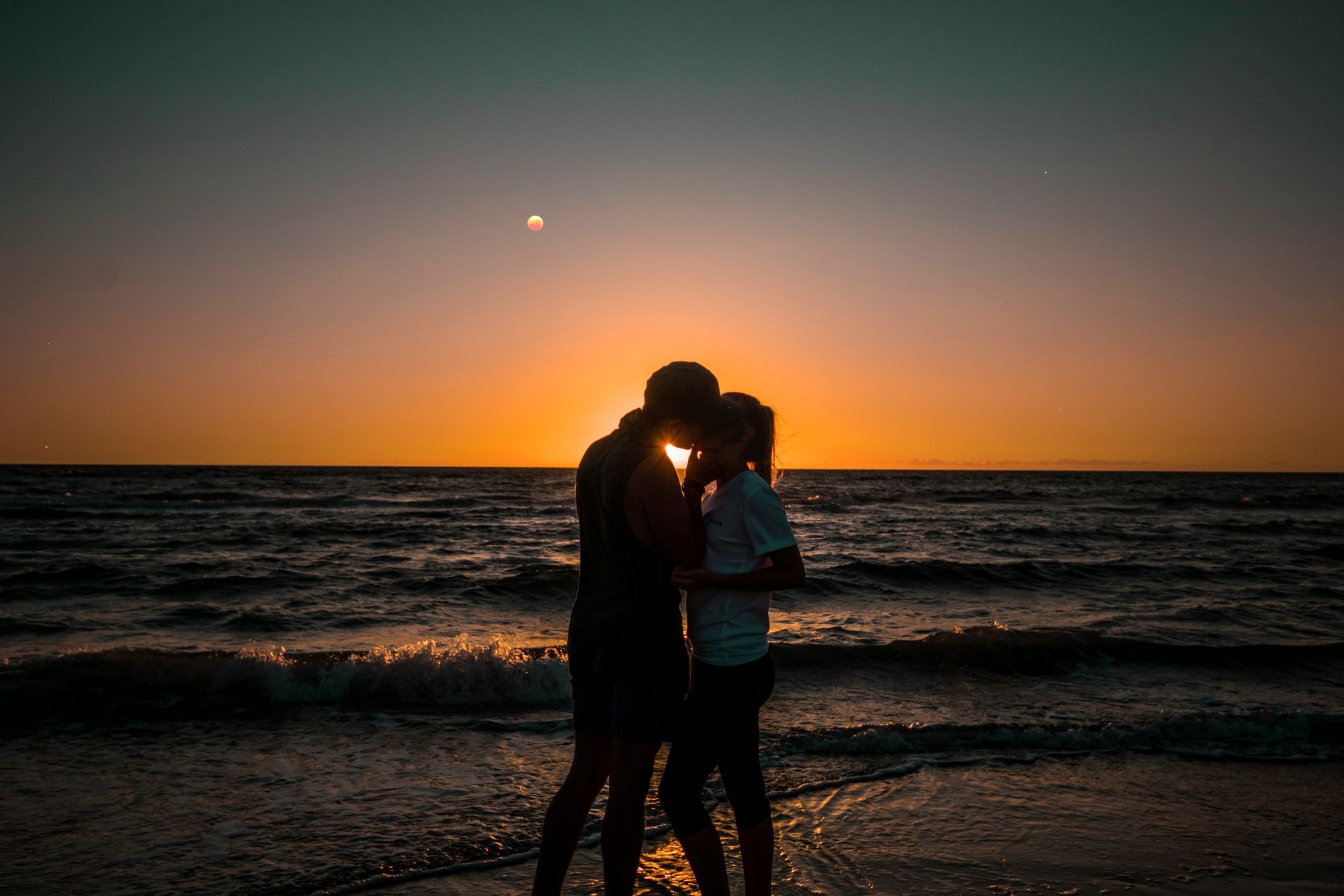 A couple on a beach at sunset looking at the moon