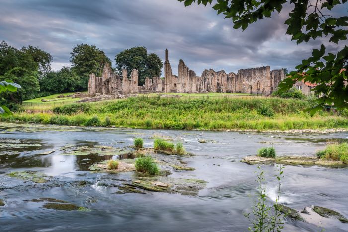 Finchale Priory ruins viewed from over a river, with green grass surrounding the stone ruins and water in the foreground.
