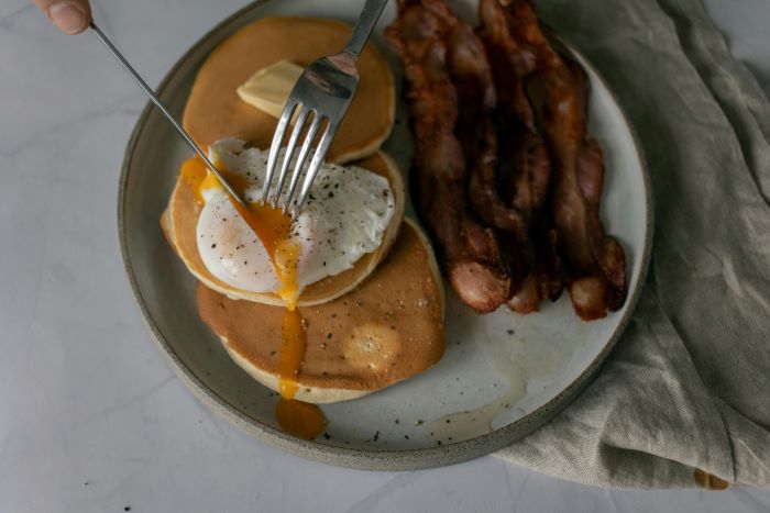 Person cutting a poached egg over some pancakes and bacon.